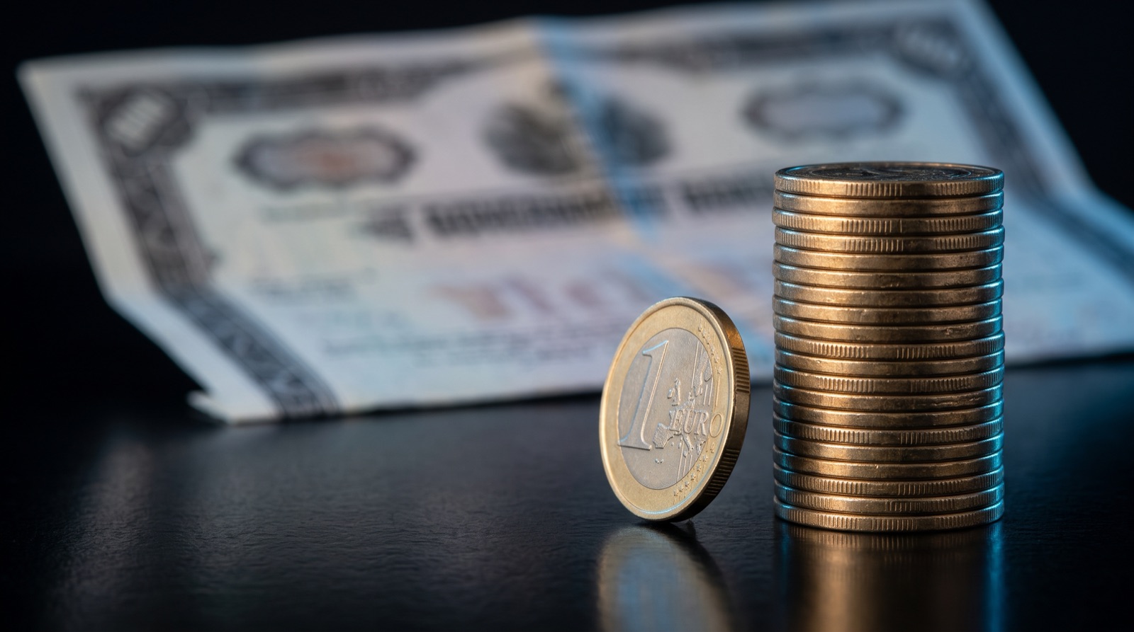 Une pièce d’un euro posée sur la tranche, légèrement inclinée, à côté d’une haute pile de pièces sur un bureau noir laqué, devant un certificat obligataire flou, sous un éclairage bleu froid